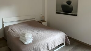 Ein ordentlich bezogenes Bett mit weißer Tagesdecke und gefalteten Handtüchern, ein Nachttisch mit Lampe sowie ein Wandbild.