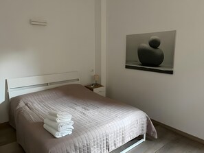 Ein ordentlich bezogenes Bett mit weißer Tagesdecke und gefalteten Handtüchern, ein Nachttisch mit Lampe sowie ein Wandbild.