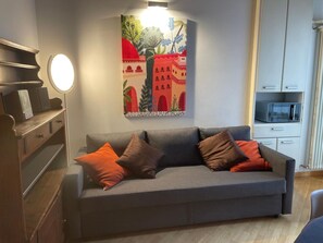 Ein Wohnzimmer mit grauem Sofa, orangefarbenen und braunen Kissen, einem Wandbild, einem runden Spiegel, einem weißen Schrank mit Mikrowelle und einem Holzschrank mit Flachbildfernseher.