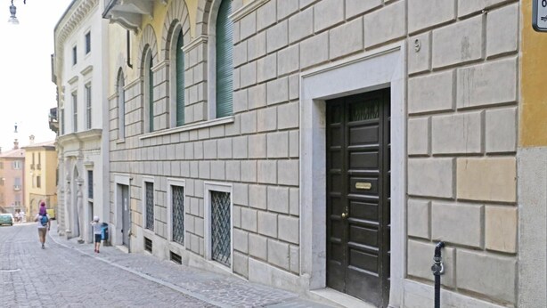Une rue pavée bordée de bâtiments historiques, une porte métallique fermée et une petite fenêtre ornée d’une grille en métal.