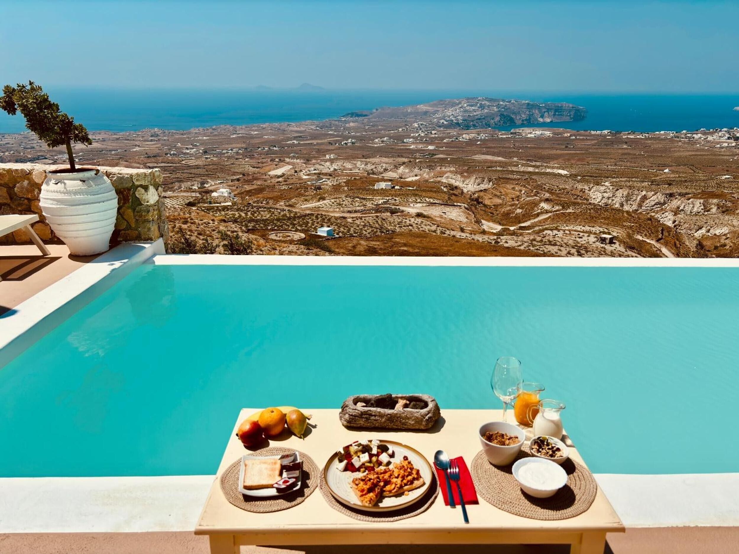 Ein Tisch am Pool mit Frühstücksangeboten, Blick auf eine Küstenlandschaft und eine Steinmauer.