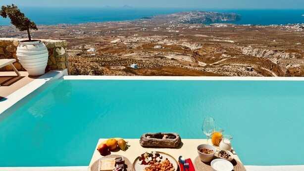 Une table placée au bord de la piscine, avec des aliments pour le petit-déjeuner, une vue sur un paysage côtier et un mur en pierre.