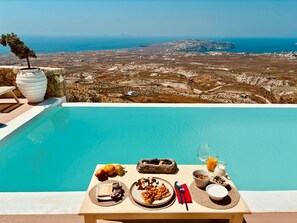 Une table placée au bord de la piscine, avec des aliments pour le petit-déjeuner, une vue sur un paysage côtier et un mur en pierre.