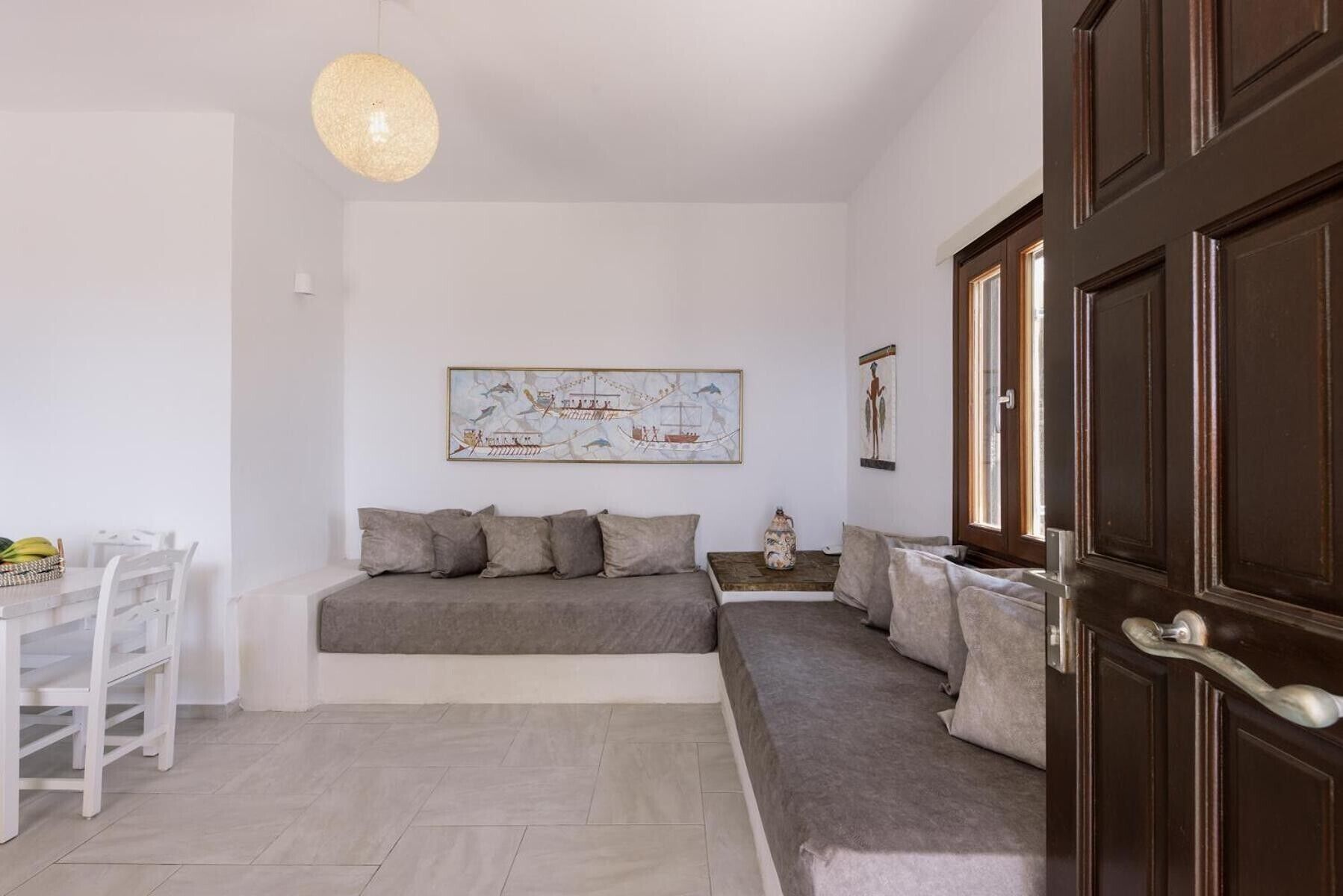 Una habitación con una cama, una mesa de comedor y una silla. Hay un cuadro en la pared y una puerta con pomo.