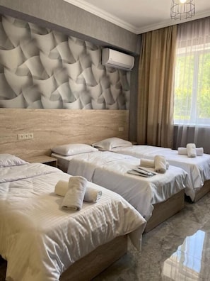 Hotellihuone, jossa on kolme sänkyä, moderni sängynpääty ja suuri ikkuna, jossa on verhot.