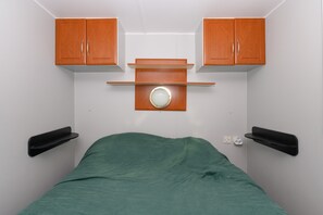 Ein kleines Zimmer mit einem Einzelbett, einer grünen Tagesdecke, Holzschränken und einem an der Wand befestigten Regal mit einem Spiegel.