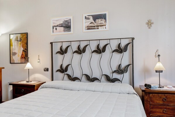 A bedroom with a bed, two bedside tables with lamps, a decorative metal headboard, and two framed pictures on the wall.