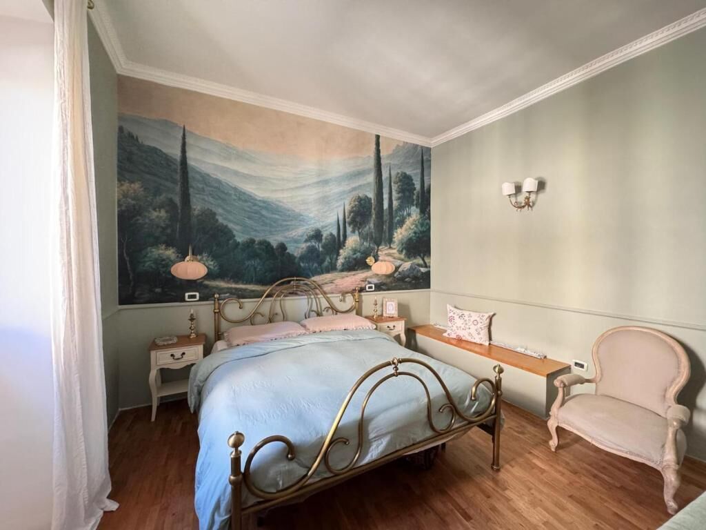 A bedroom with a bed, a nightstand, a chair, a wall-mounted lamp, and a large landscape painting.