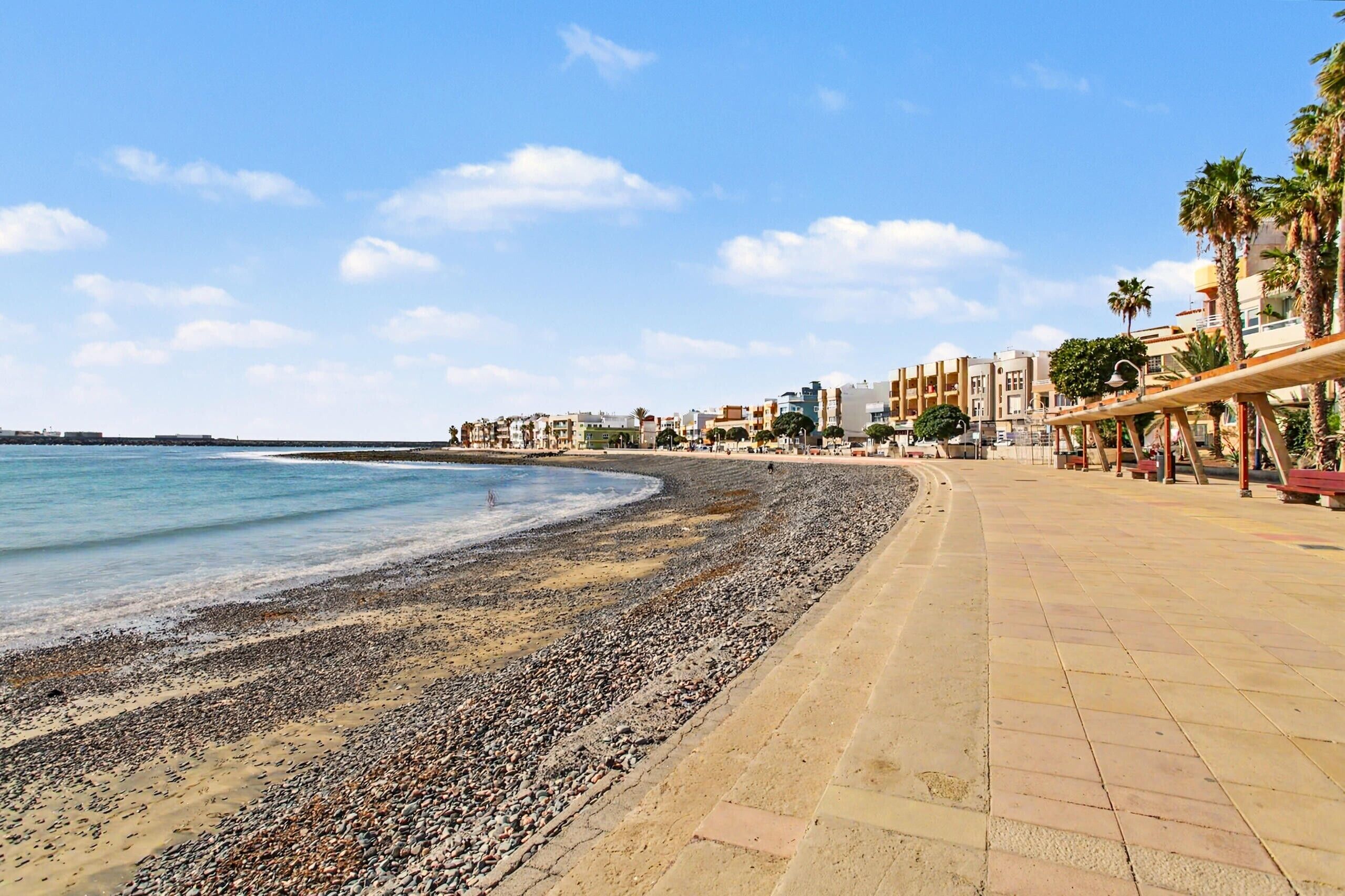 Una playa de guijarros, un camino de madera y edificios al fondo.