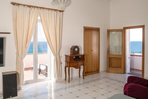 Una habitación con un ventanal grande, una puerta de madera, una mesa pequeña con un reloj, una silla roja y un altavoz.