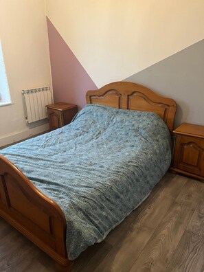 Una cama individual con estructura de madera y colcha estampada, una mesita de noche, un pequeño armario de madera y un radiador en la pared.
