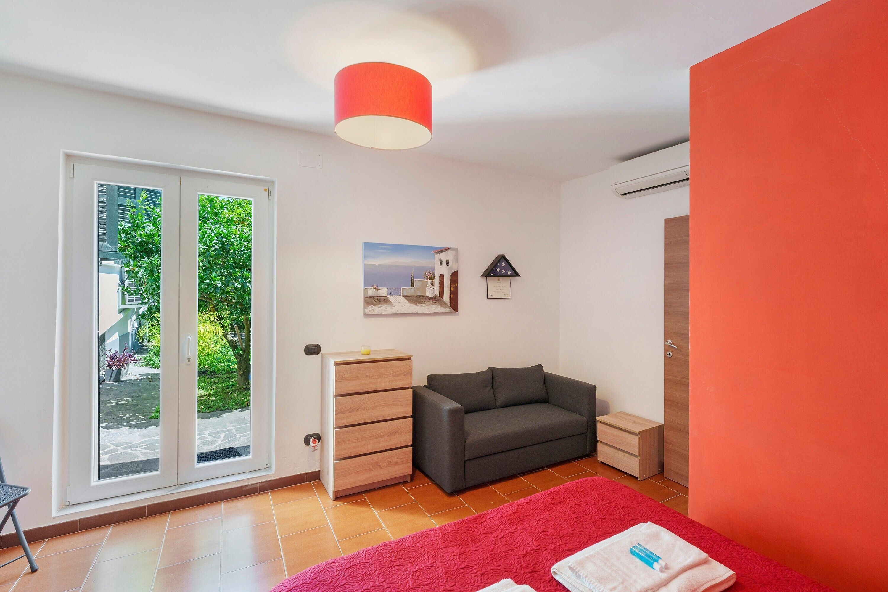 Una camera da letto con una porta scorrevole in vetro che dà su un balcone, un divano grigio, un comò di legno, un tappeto rosso e una parete rossa.