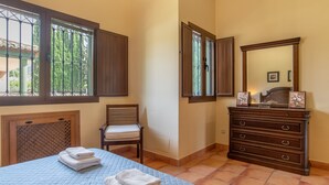 Ein Schlafzimmer mit Bett, Stuhl, Kommode, Spiegel und einem Fenster mit Blick auf Bäume.