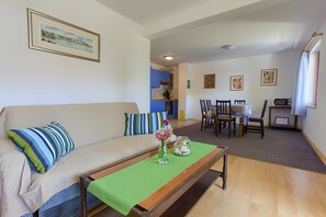 Uma sala de estar com um sofá, uma mesa de jantar com cadeiras, uma cozinha ao fundo e um quadro na parede.