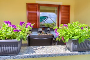 An outdoor dining area with a table set for two, surrounded by potted plants with purple flowers, and a view through a window with wooden shutters.