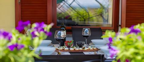Mesa posta com taças de vinho, uma garrafa e aperitivos, com vista para o verde através de uma janela.