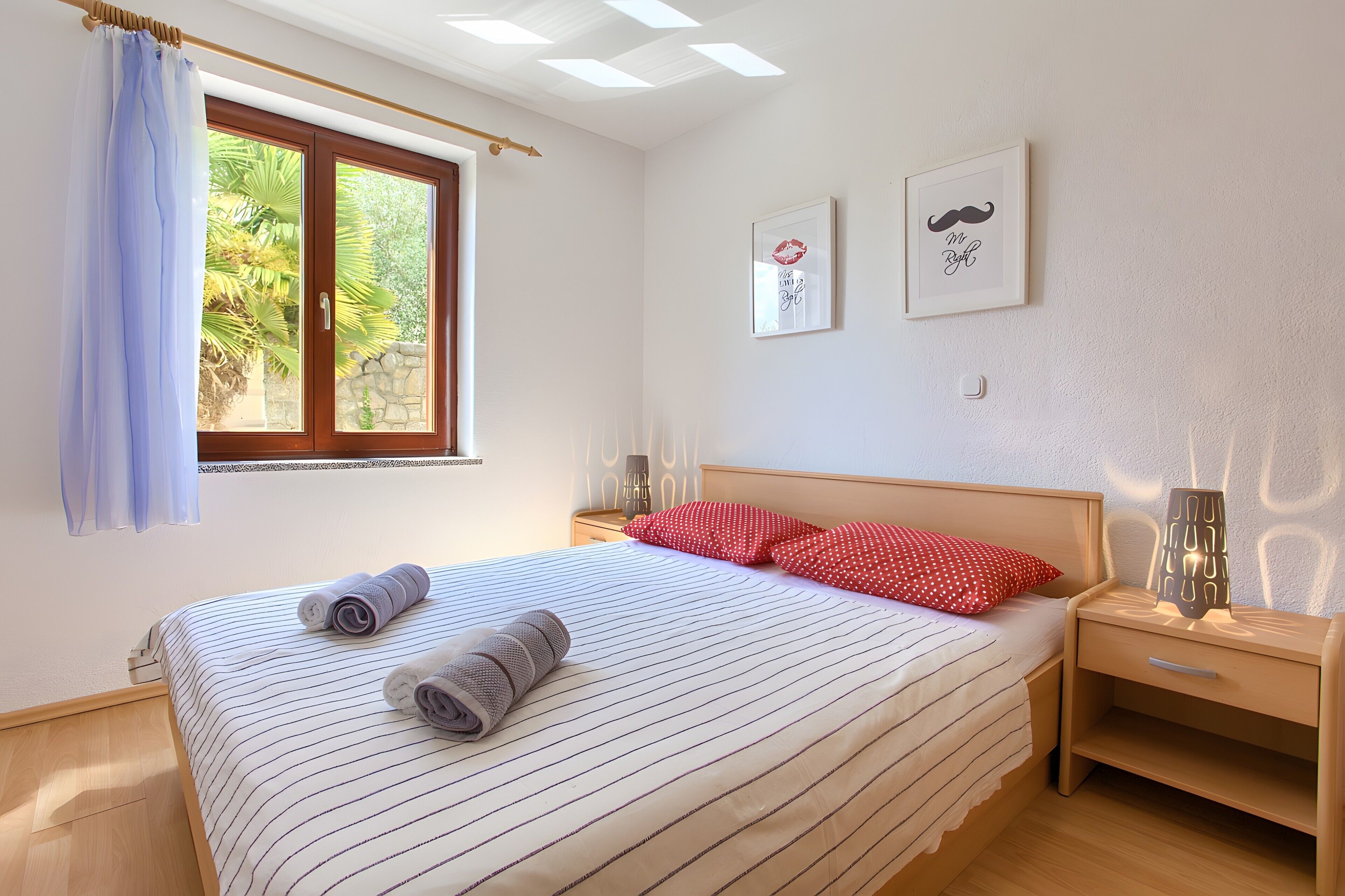 Una cama bien hecha con ropa de cama a rayas, una ventana con cortinas, una mesita de noche con lámpara y dos cuadros en la pared.