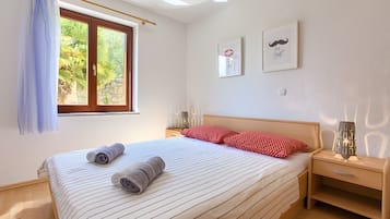 Una cama bien hecha con ropa de cama a rayas, una ventana con cortinas, una mesita de noche con lámpara y dos cuadros en la pared.
