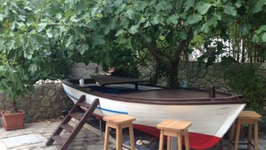 Un bateau avec un pont en bois, un banc et des tabourets, amarré près d’un mur de pierre et sous un arbre.