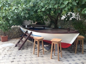 Um barco com convés de madeira, um banco e banquinhos, atracado perto de um muro de pedra e sob uma árvore.