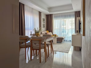 A modern living room with a dining area, large windows, and a flat-screen TV. - Qoople Green Ocean B55 2-bedroom apartment in serene Alanya (Alanya)