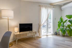 Un salon moderne avec un téléviseur à écran plat posé sur une console en bois, un lampadaire, une grande plante en pot et une porte-fenêtre coulissante donnant sur un espace extérieur.