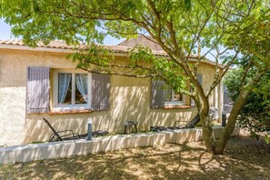 Une maison avec un toit en tuiles, deux fenêtres avec volets, une petite table et une chaise à l’extérieur.