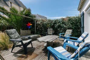 Um pátio externo com piso de madeira, uma mesa de madeira e duas cadeiras azuis com almofadas listradas.