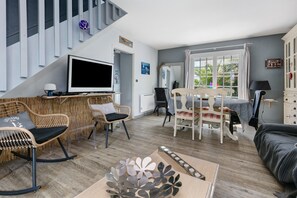 Ett modernt vardagsrum med en platt-TV, möbler i rotting, ett matbord med stolar och en trappa.