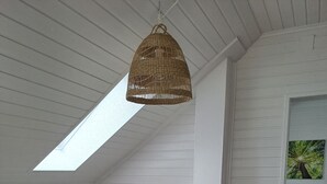 A dining area with a wooden table and chairs, a woven pendant light, a skylight, and a view of greenery outside.