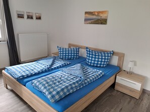 Ein ordentlich bezogenes Bett mit blauer Bettwäsche, ein Holz-Kopfteil, ein Nachttisch mit Lampe und eine Wand mit drei kleinen Bildern.