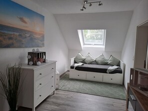 Um quarto com teto inclinado, uma janela, cama com almofadas, cómoda e um quadro na parede.