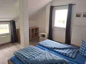 Quarto com teto inclinado, janela com cortinas, pequena mesa de cabeceira com uma planta e um móvel de armazenamento em madeira.