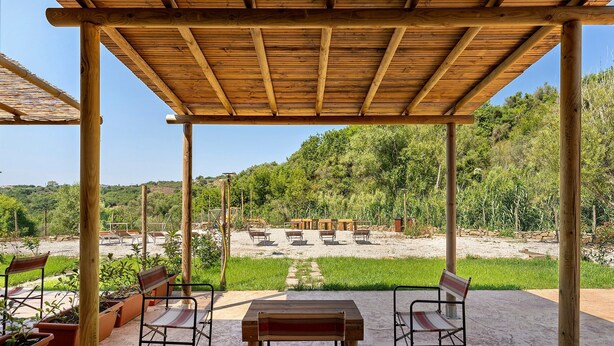Un patio al aire libre con mesas y sillas de madera, rodeado de follaje y un cielo despejado.