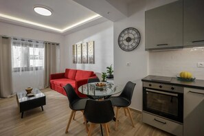 A modern kitchen with a built-in oven, a dining area with a glass table and chairs, a red sofa, and a wall clock. - Apartment 'Polisher' with Shared Terrace, Wi-Fi and Air Conditioning (Heraklion)