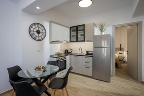 Una cocina compacta con armarios blancos, un refrigerador de acero inoxidable, un reloj redondo y un comedor con una mesa de vidrio y sillas negras.