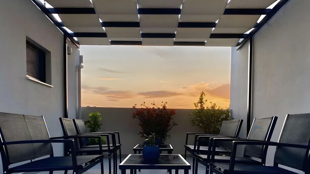 Una terraza en la azotea con vistas a la puesta de sol, con una mesa, sillas, plantas en macetas y una ventana pequeña.