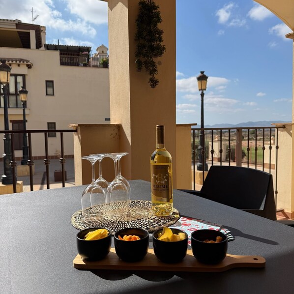 Uma mesa com uma garrafa de vinho Blume, três copos de vinho e quatro recipientes com petiscos numa varanda com vista para edifícios e um céu claro.