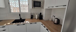 A modern kitchen with white cabinets, a black sink, a microwave, a kettle, and a window with a decorative screen.