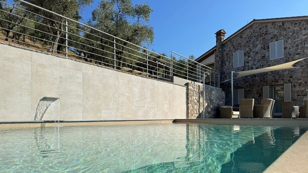 Una piscina cristalina con un muro de piedra, mobiliario exterior y un edificio de piedra al fondo.