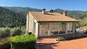 Un edificio de piedra con techo de tejas, grandes ventanales y vistas a una cadena montañosa boscosa.