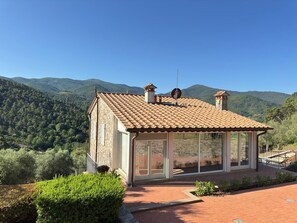 A stone building with a tiled roof, large glass windows, and a view of a forested mountain range.