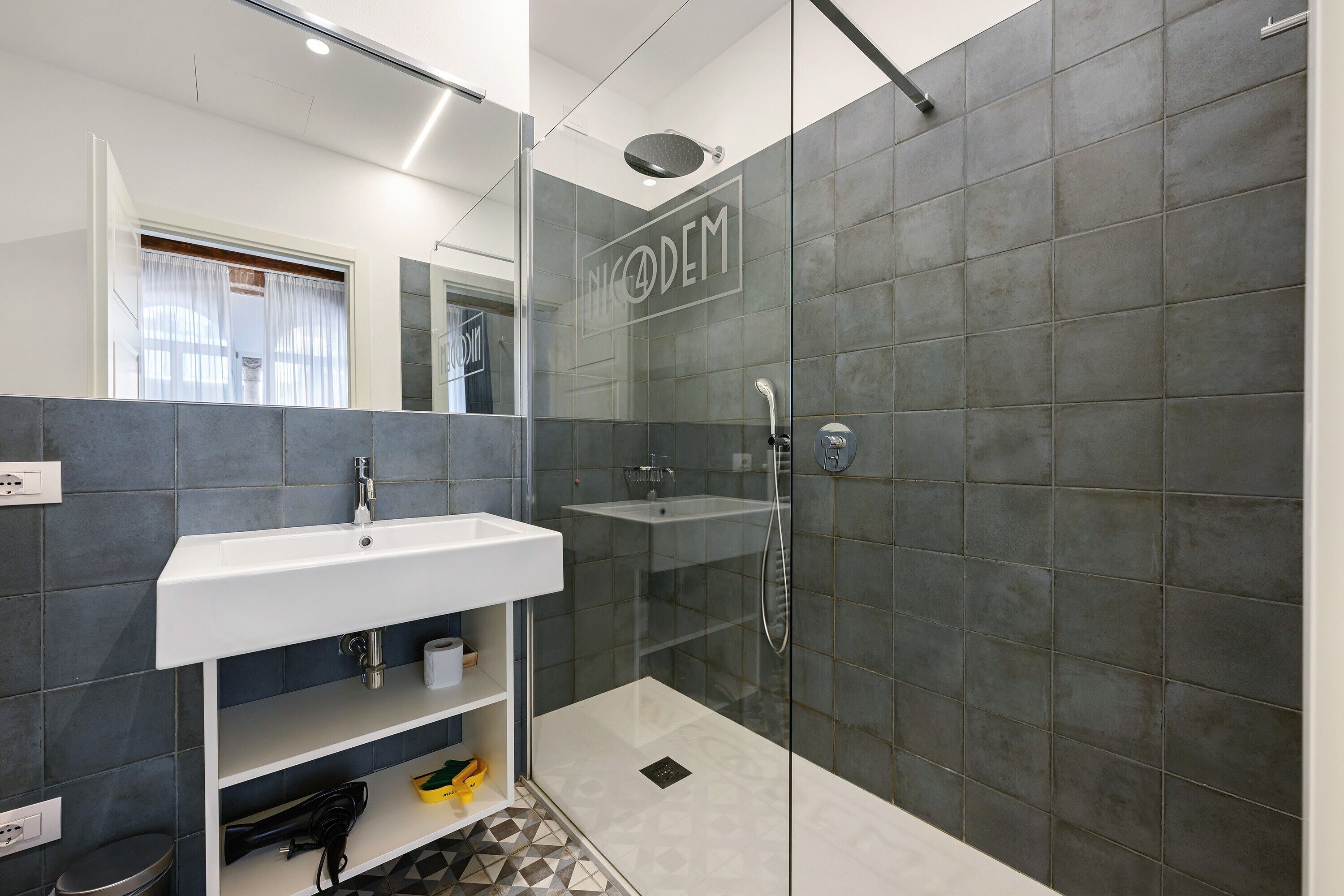 Un baño moderno con cabina de ducha de vidrio, un lavabo blanco y paredes cubiertas de baldosas oscuras.