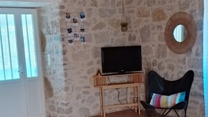A spacious room with a stone wall, a wooden ceiling, a television, a black chair, a red stool, and a tiled floor. - Le gite de L' Alauzet – Charming Stone House with Panoramic Tarn Views (Paulhe)