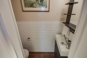 Une salle de bain avec des murs recouverts de carreaux blancs, des toilettes, un lavabo avec un robinet et un tableau encadré accroché au mur.