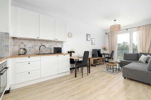 A modern kitchen with white cabinets, a dining area with a table and chairs, a gray sofa, and a large window with curtains. - Modern Apartment with a Balcony | A Place to Work (Chorzów)