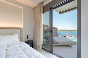 Una habitación de hotel moderna con cama, una mesita de noche pequeña, una puerta de cristal que da a un balcón con piscina y sillas de descanso.