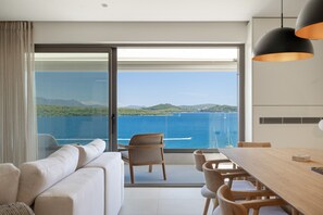 Ein modernes Zimmer mit einer großen Schiebeglasdoor, die auf einen Balkon mit Meerblick führt. Es gibt ein Sofa, einen Esstisch mit Stühlen und eine Pendelleuchte.