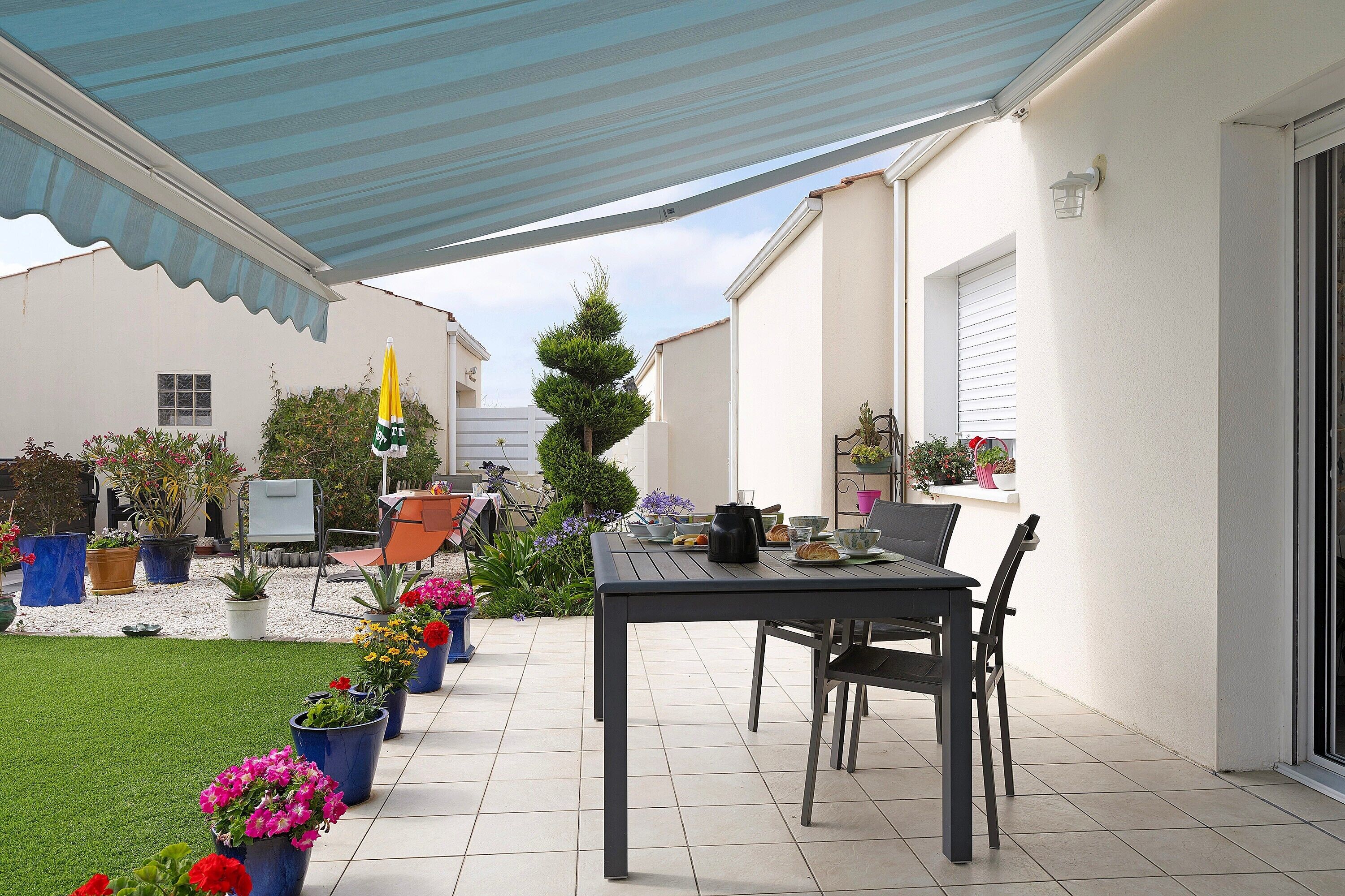 A patio area with a table and chairs, surrounded by potted plants and a blue awning.