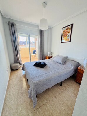 A bedroom with a bed, a bedside table, a lamp, a chair, a window with curtains, and a painting on the wall.